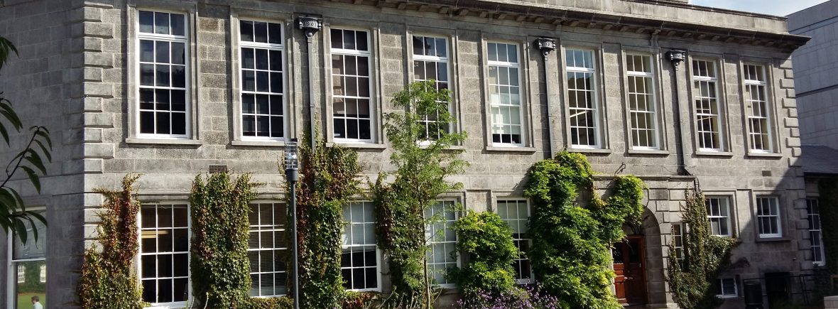 The Botany Department at Trinity College Dublin. The early 20th century building has flower beds in front with climbing plants growing up the walls. There are benches in front of the building.