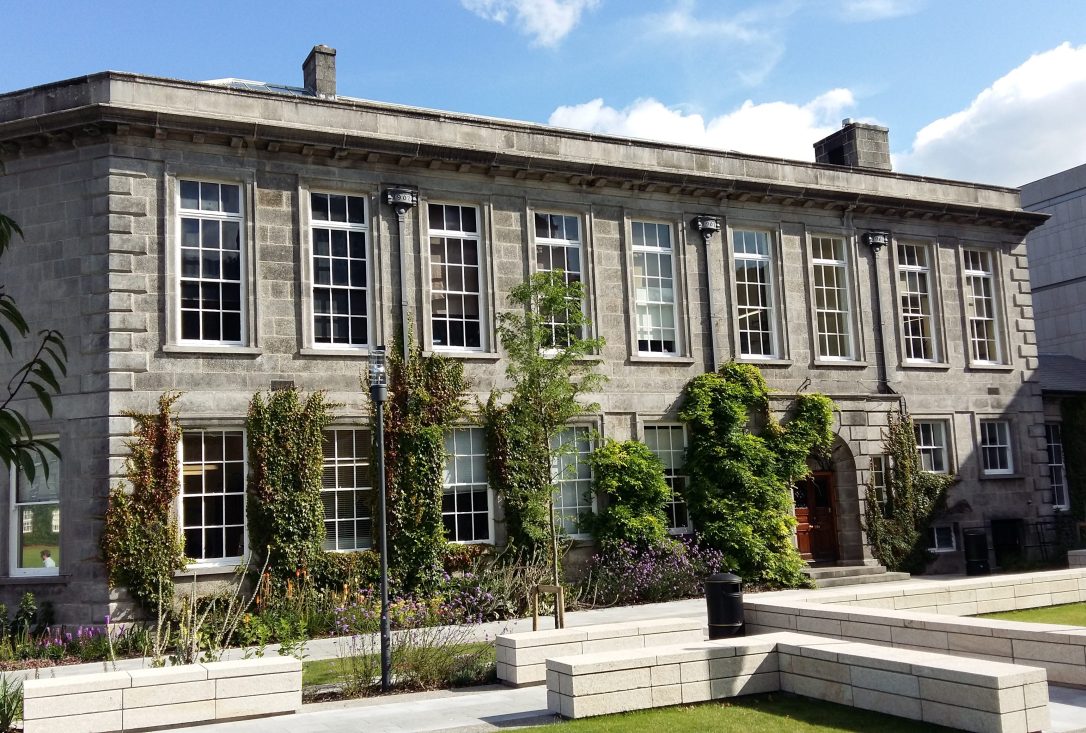 The Botany Department at Trinity College Dublin. The early 20th century building has flower beds in front with climbing plants growing up the walls. There are benches in front of the building.